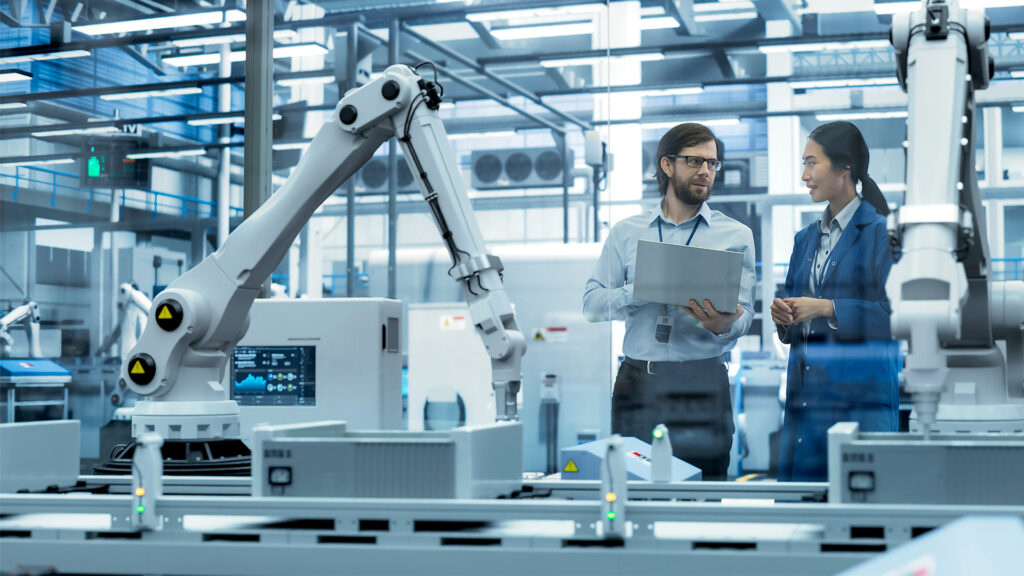 Young Engineer and Asian Technician Having a Conversation and Using Laptop Computer. Specialists Monitoring, Analyzing Conditions at a Modern Electronics Factory with Automated Assembly Line
